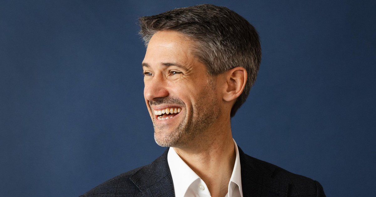 A man with short gray hair and a beard is smiling broadly, looking to his left, wearing a white shirt and a dark blazer against a blue background.