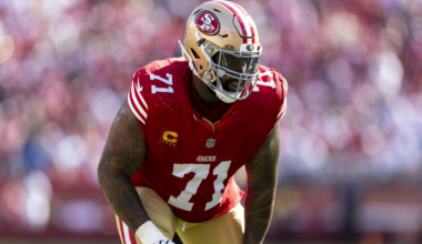 A San Francisco 49ers football player wearing a red jersey with number 71 and a gold helmet is poised in a defensive stance on a green field.