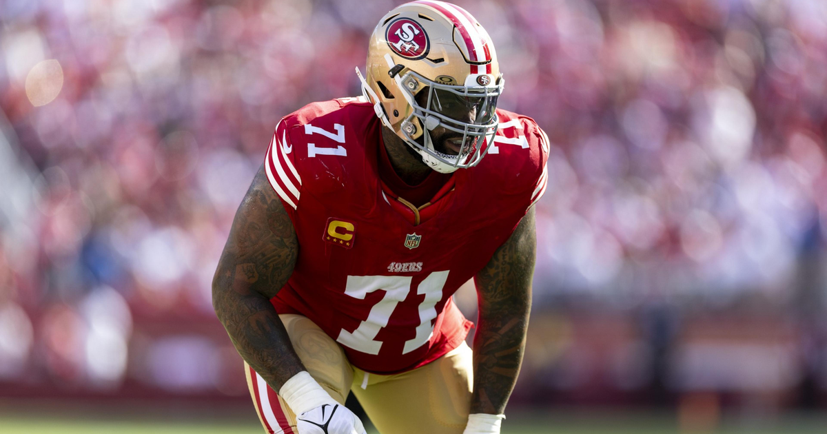 A San Francisco 49ers football player wearing a red jersey with number 71 and a gold helmet is poised in a defensive stance on a green field.