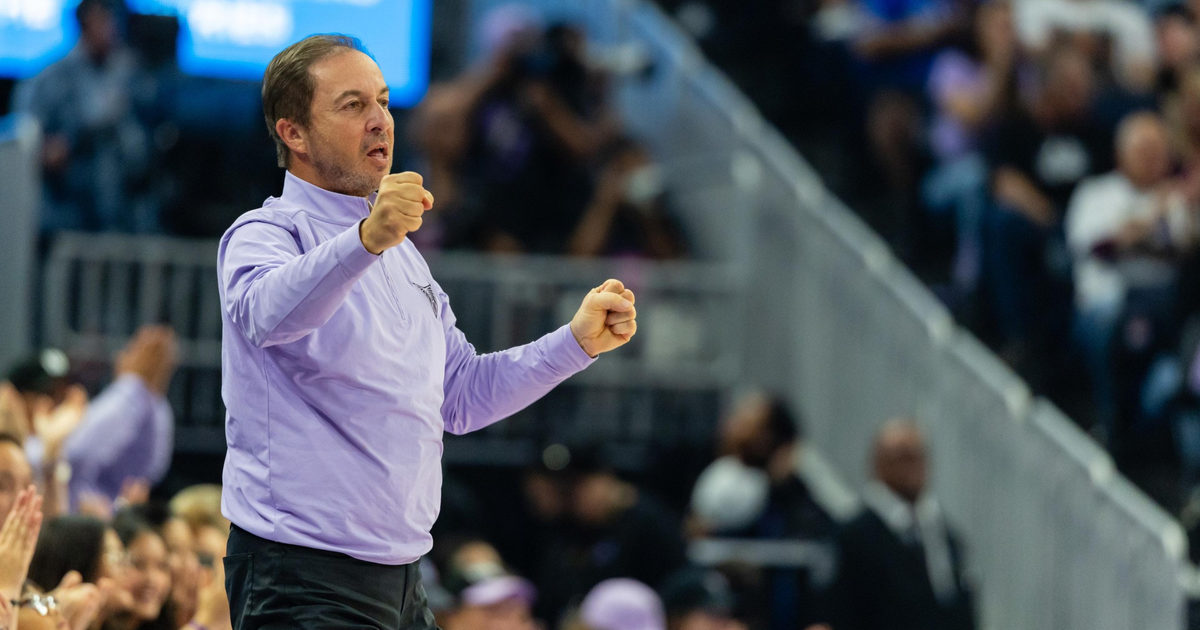 A man in a light purple shirt and black pants clenches his fists and appears to be shouting, standing in front of a cheering crowd.