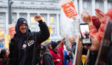 A crowd of people, some holding signs reading "ON STRIKE FOR SPECI STUDENTS EDUCATORS," gathers outdoors, with one man raising his fist and smiling.
