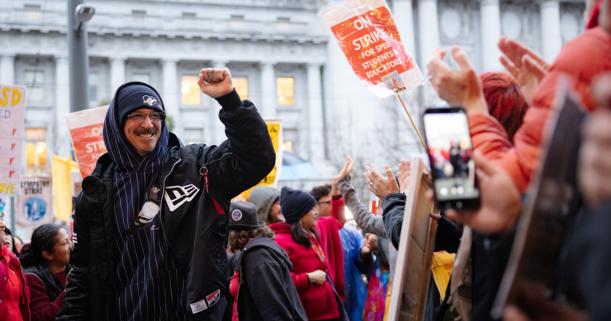 A crowd of people, some holding signs reading "ON STRIKE FOR SPECI STUDENTS EDUCATORS," gathers outdoors, with one man raising his fist and smiling.