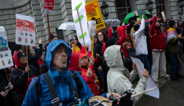 A group of protesters in rain gear hold signs saying "ON STRIKE" while one person plays a drum, standing in front of a stone building with arched windows.