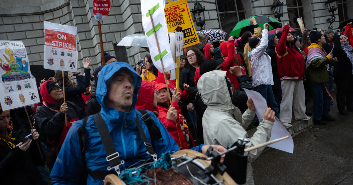 A group of protesters in rain gear hold signs saying "ON STRIKE" while one person plays a drum, standing in front of a stone building with arched windows.