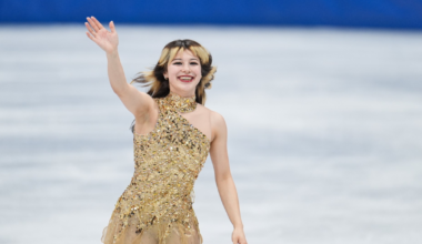 A female figure skater in a sparkling gold dress smiles and waves with her right hand while standing on the ice rink.