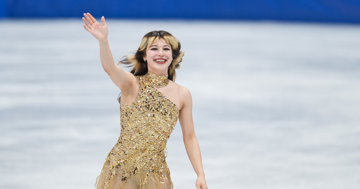 A female figure skater in a sparkling gold dress smiles and waves with her right hand while standing on the ice rink.