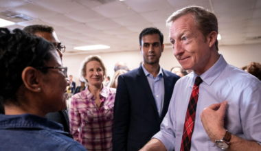 A man in a white shirt and red plaid tie speaks to a group of people, including a woman in glasses and a plaid shirt, in a room with a drop ceiling.