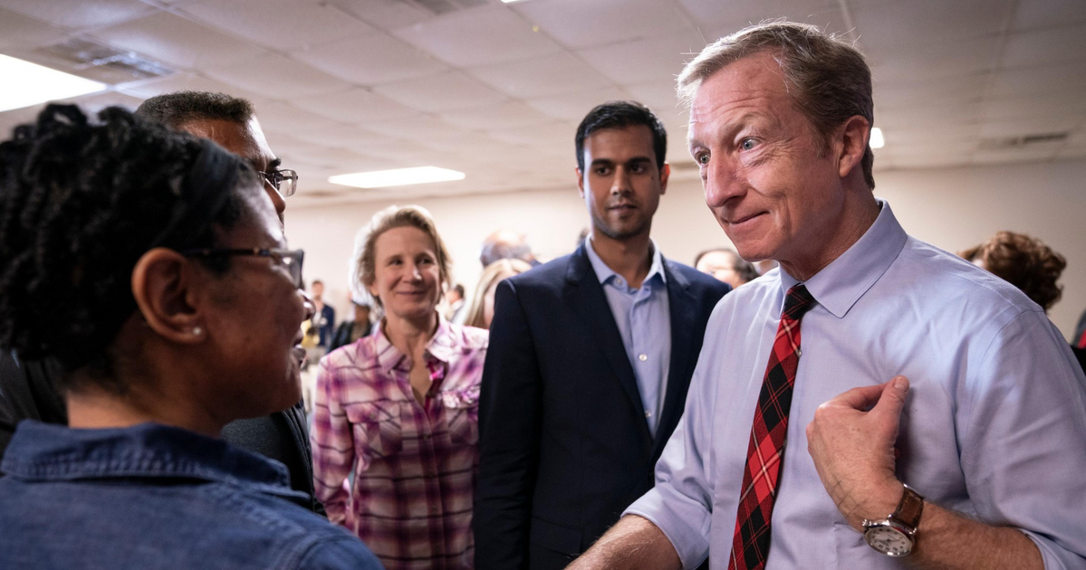 A man in a white shirt and red plaid tie speaks to a group of people, including a woman in glasses and a plaid shirt, in a room with a drop ceiling.