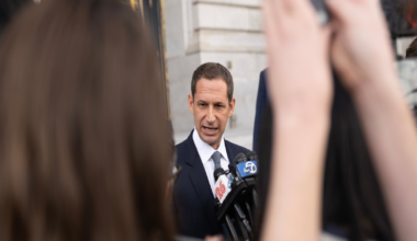 A man in a dark suit speaks into multiple microphones as several people hold up phones and cameras in front of him.