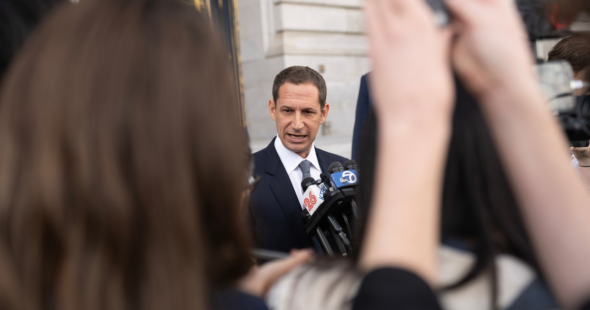A man in a dark suit speaks into multiple microphones as several people hold up phones and cameras in front of him.