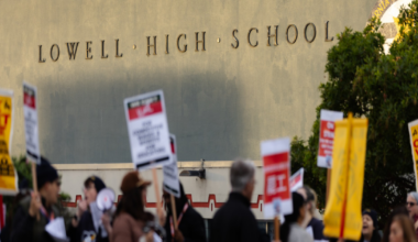 People holding protest signs gather outside a building labeled "Lowell High School" under a clear sky.