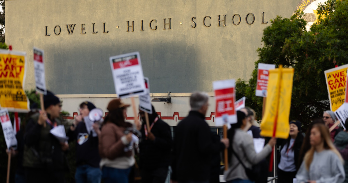 People holding protest signs gather outside a building labeled "Lowell High School" under a clear sky.