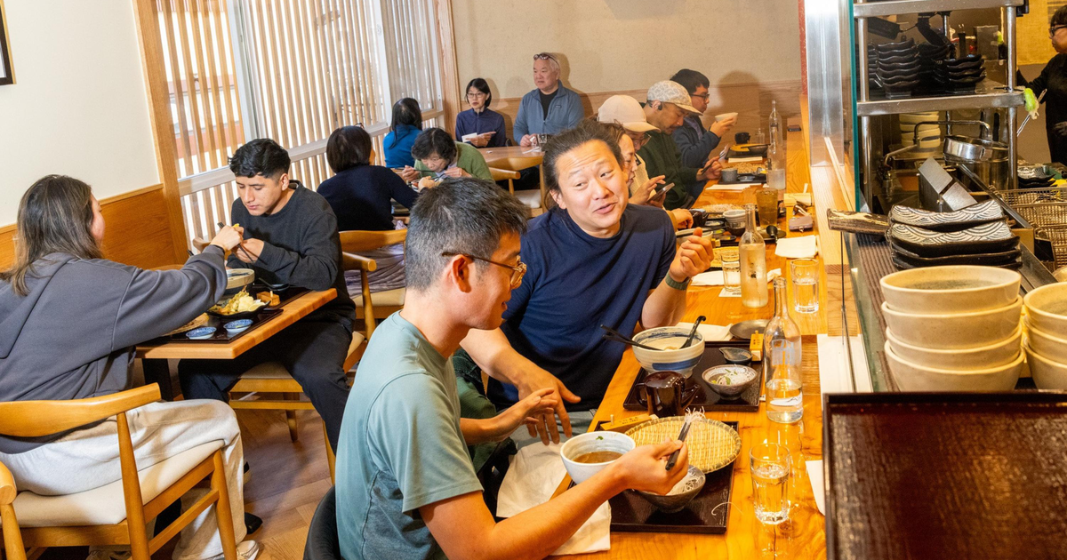 Three people are seated at wooden tables in a restaurant, eating with chopsticks while a server places a dish on the table.