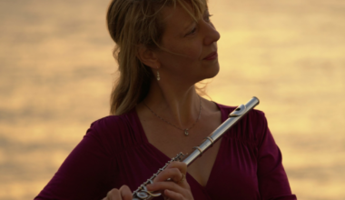 A woman in a purple top holds a silver flute near her chest, gazing thoughtfully toward the right, with a warm, blurred sunset background.