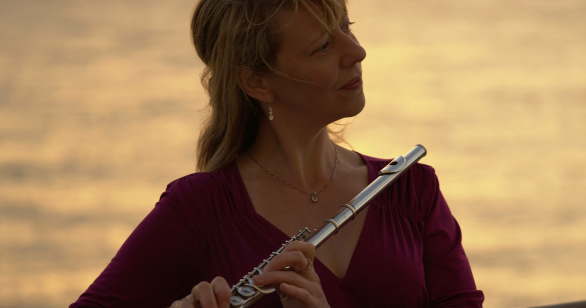 A woman in a purple top holds a silver flute near her chest, gazing thoughtfully toward the right, with a warm, blurred sunset background.