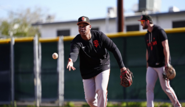 A baseball player in San Francisco Giants gear is throwing a ball on a dirt field while another player stands in the background holding a glove.