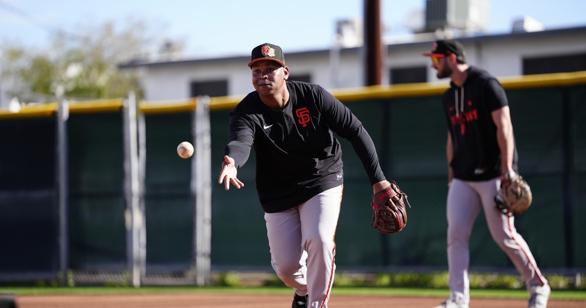 A baseball player in San Francisco Giants gear is throwing a ball on a dirt field while another player stands in the background holding a glove.