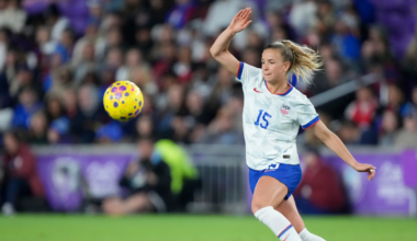A female soccer player wearing a white USA jersey with number 15 kicks a yellow and purple ball on a green field with blurred spectators in the background.