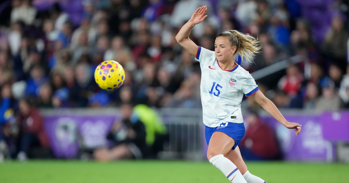 A female soccer player wearing a white USA jersey with number 15 kicks a yellow and purple ball on a green field with blurred spectators in the background.
