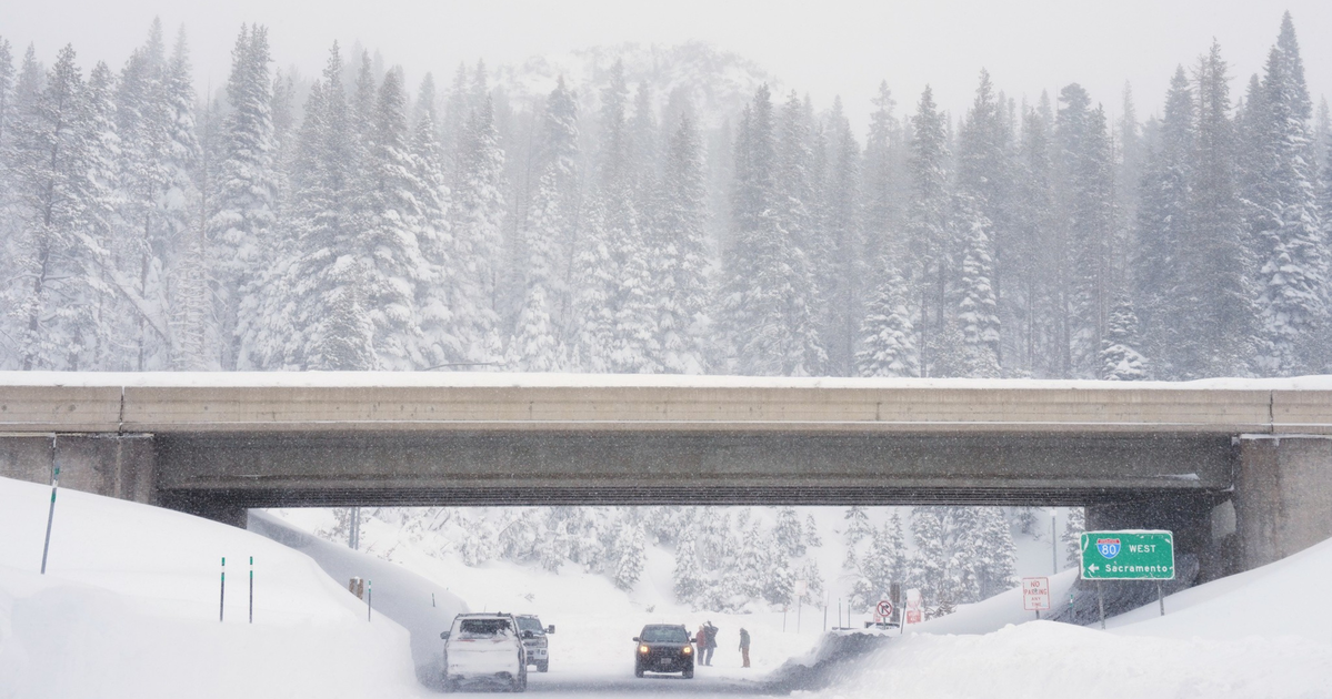 A snowy highway under a bridge with cars and people, surrounded by snow-covered trees and thick snow on the ground.