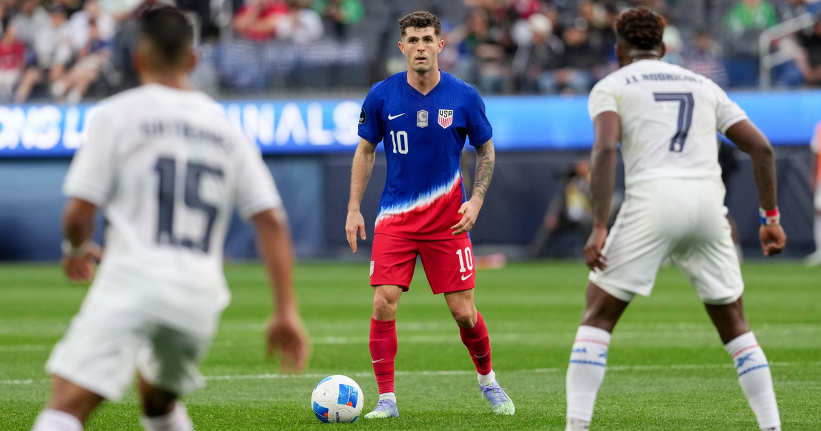 A soccer player in a blue and red US uniform stands poised with the ball, facing two opposing players in white uniforms.