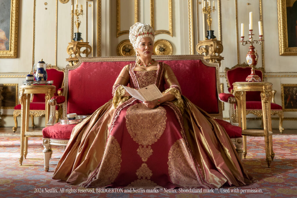 Her Majesty the Queen on Bridgerton sitting on a red sofa.