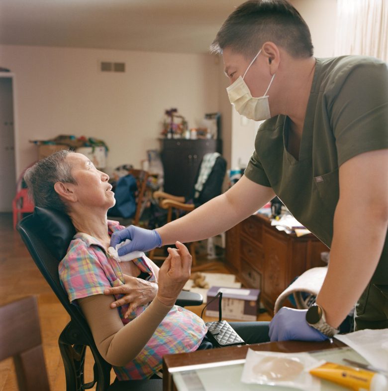 A caregiver wearing a mask and gloves tends to an elderly woman seated in a wheelchair at home. The scene conveys care and support in a cozy setting.