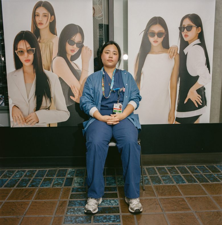 A person in blue scrubs sits on a chair in front of a fashion poster featuring stylish models. The scene conveys contrast and casualness.