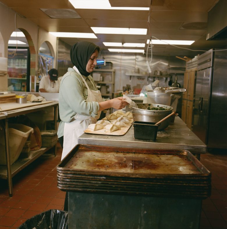 A woman in a headscarf and apron prepares pastries in an industrial kitchen. She fills dough with stuffing, conveying focus and culinary skill.