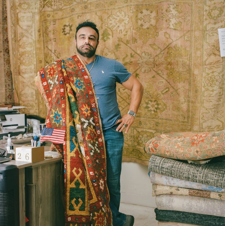 Man in a blue shirt stands confidently, draped with a vibrant red patterned rug. Background of stacked, elaborate rugs and a small desk with an American flag.