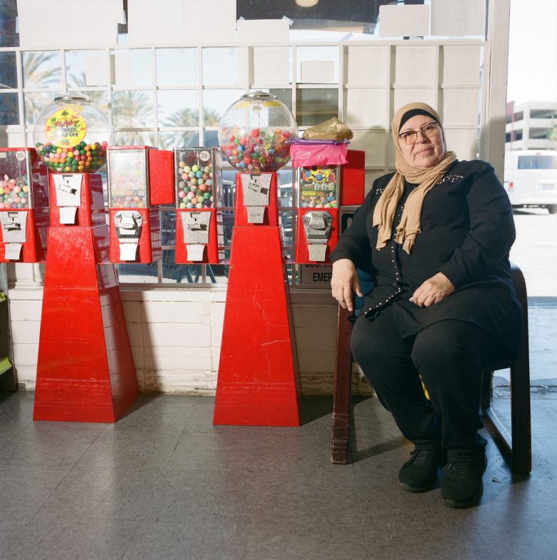A woman in a hijab sits beside bright red gumball machines, exuding a calm and content demeanor. Sunlight filters through a window, creating a cozy atmosphere.