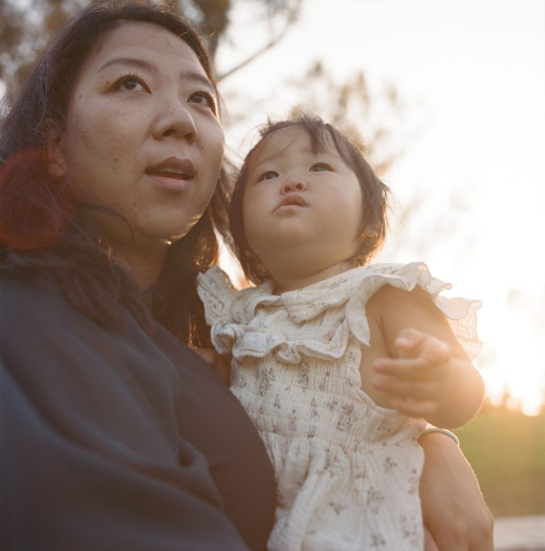 A woman and a child look upward, bathed in warm sunlight. The child wears a white dress with ruffles. The scene conveys a peaceful, hopeful mood.