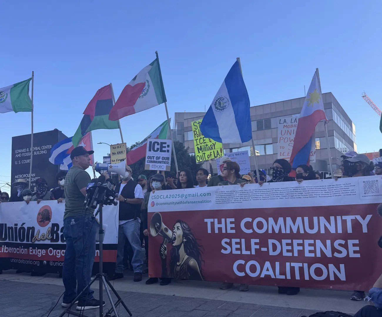A man speaks into a megaphone facing a large group of people holding flags, signs, and banners. A large banner at the front of the crowd reads "The Community Self-Defense Coalition." They are outside, with a large building behind them and clear, blue sky above.