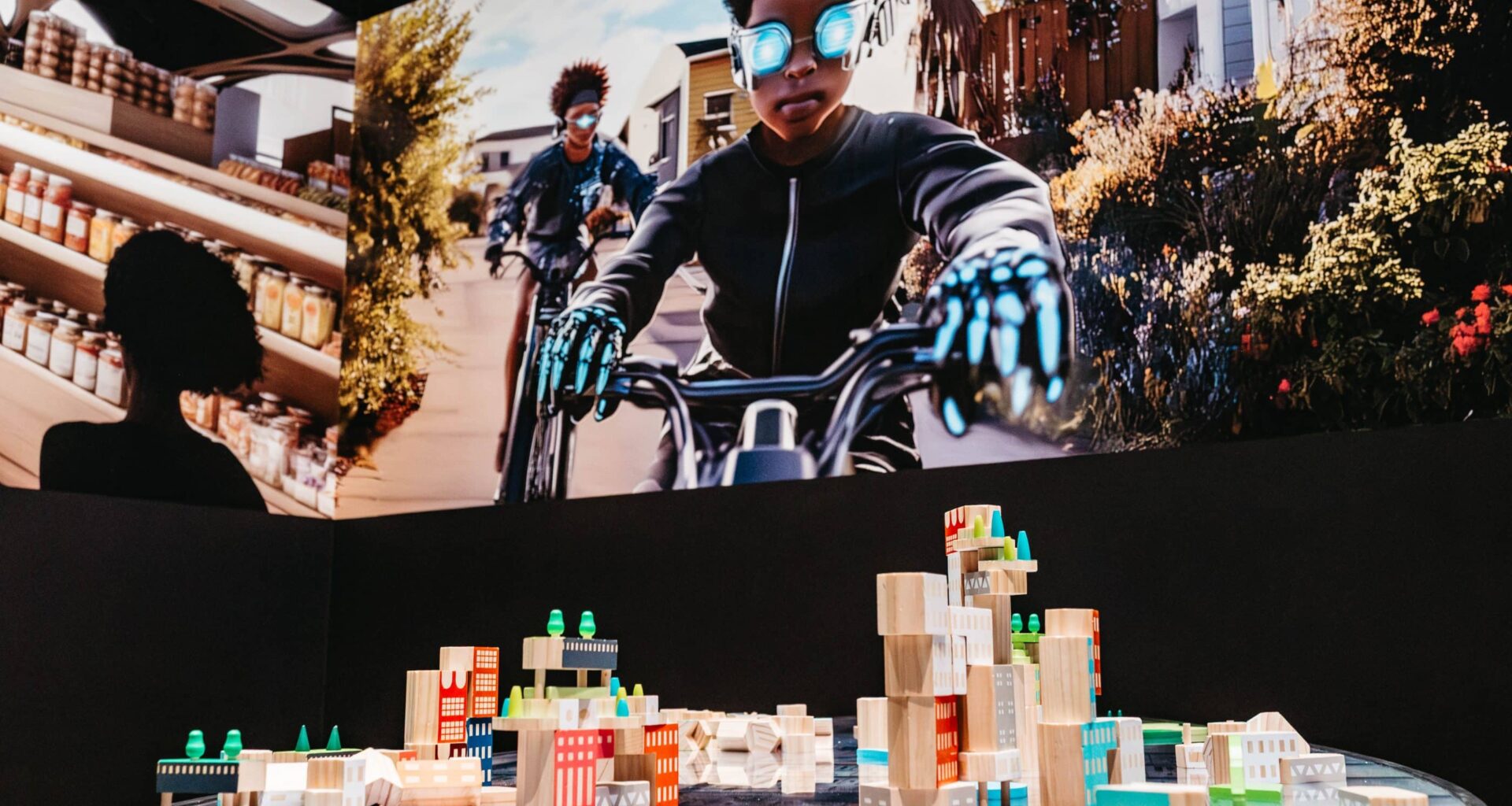 A group of building blocks on a table with buildings printed on them. Above them is a projected image of two boys on bikes wearing futuristic gear.
