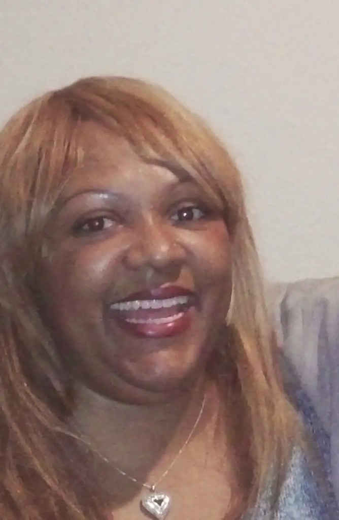 A person with long, light brown hair is smiling widely, wearing a heart-shaped necklace. The background is plain, emphasizing their joyful expression.