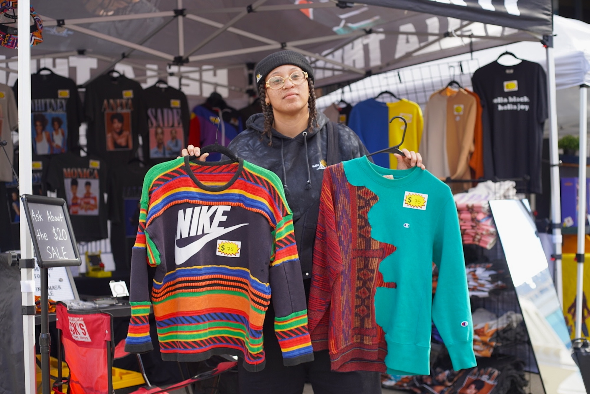 A woman in glasses and a hat holds up two handmade sweaters on hangers.