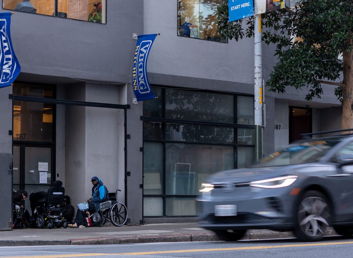 A blur of a gray car drives past a city street. A person in a wheelchair and another seated are near a building entrance with blue banners. Trees and buildings line the sidewalk.