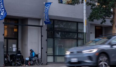 A blur of a gray car drives past a city street. A person in a wheelchair and another seated are near a building entrance with blue banners. Trees and buildings line the sidewalk.