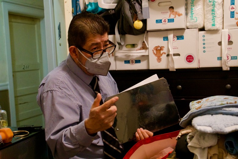 A man in an office setting with shelves of paper files is wearing a lavender button-down shirt, necktie, N-95-style face mask and glasses, and holding a stack of papers.