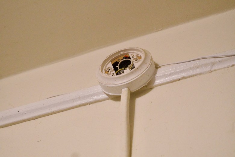 A hard-wired smoke detector placed near a ceiling has interior wires showing and appears to be missing a protective closure panel.