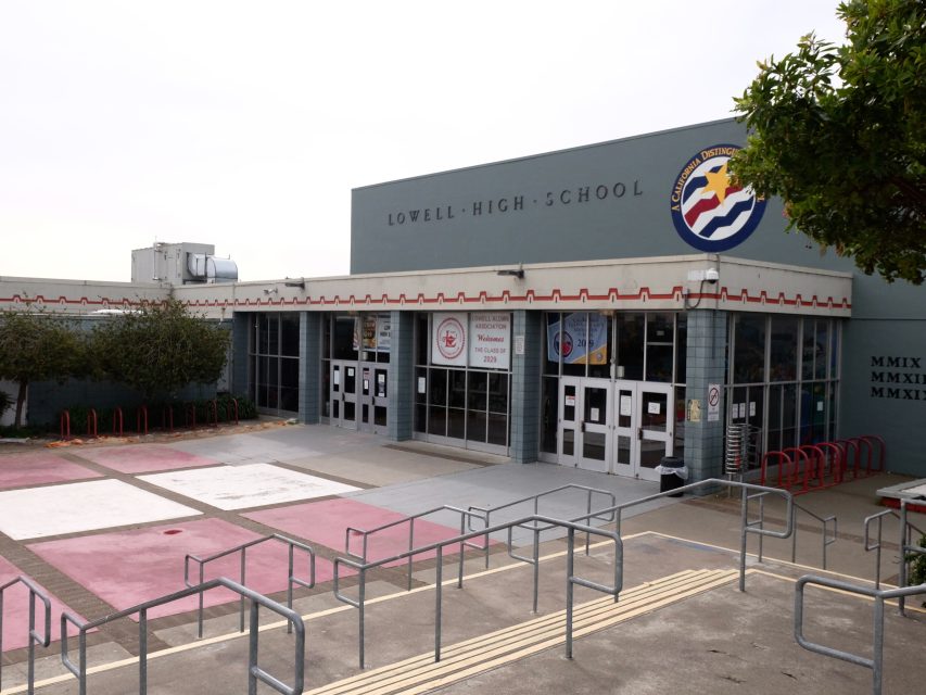 The front entrance of Lowell High School, featuring glass doors, a school logo, and an outdoor area with bike racks and railings.