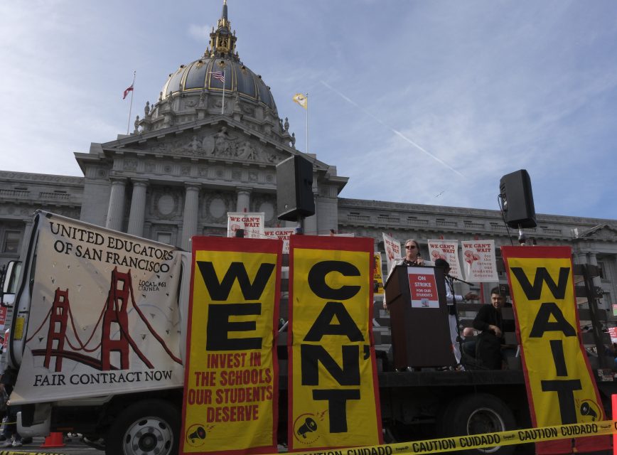 A rally in front of San Francisco City Hall features teacher-led banners reading "WE CAN'T WAIT" and "FAIR CONTRACT NOW" from the United Educators of San Francisco.