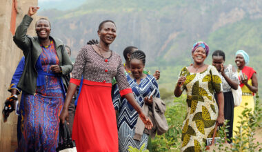 A half-dozen East African women smile and laugh as they walk by farm fields