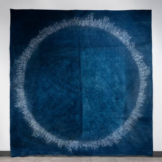 A large blue textile with a faint, circular white pattern made up of clustered script-like markings near the fabric’s edges, reminiscent of pieces found in museums, displayed against a neutral wall.