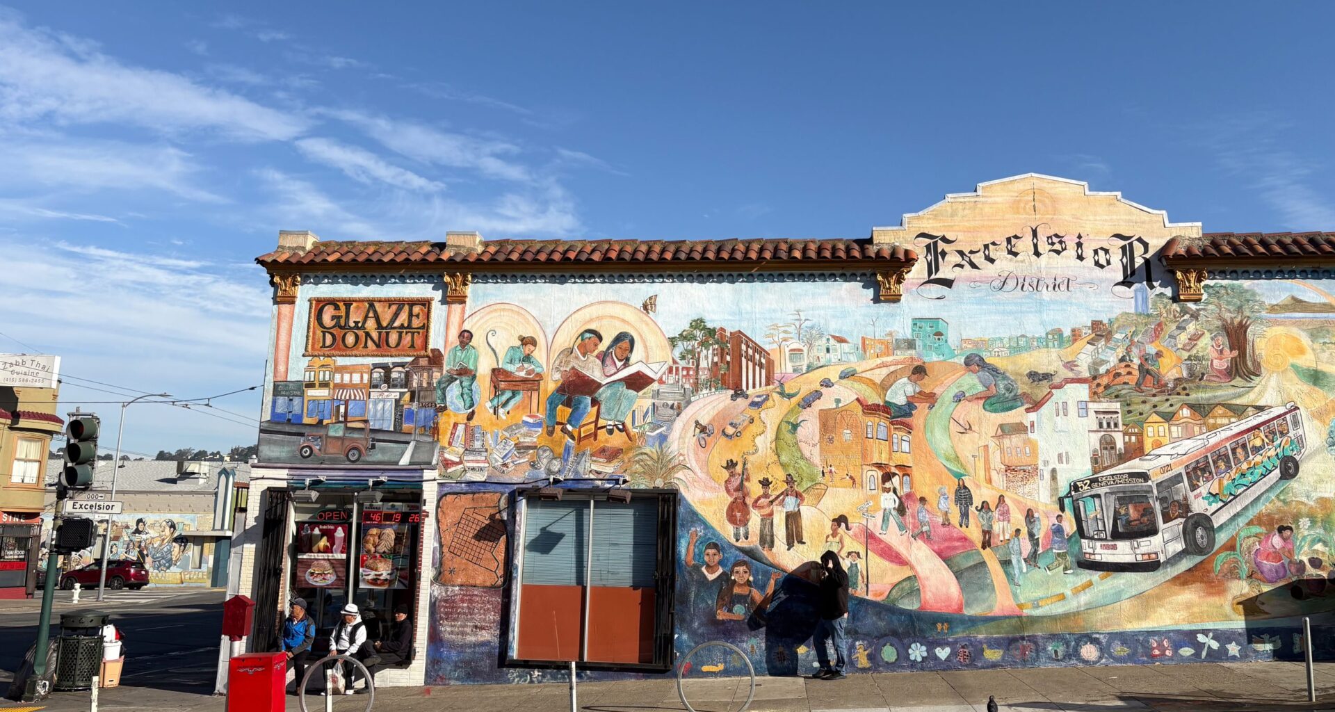 Street mural depicting diverse community scenes, including a bus and pedestrians, covers the wall of a building labeled "Excelsior District." Nearby, there's a "Glaze Donut" shop entrance.