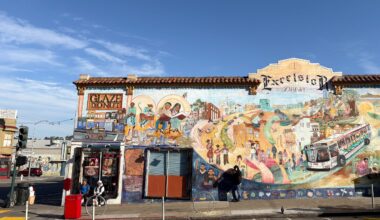 Street mural depicting diverse community scenes, including a bus and pedestrians, covers the wall of a building labeled "Excelsior District." Nearby, there's a "Glaze Donut" shop entrance.