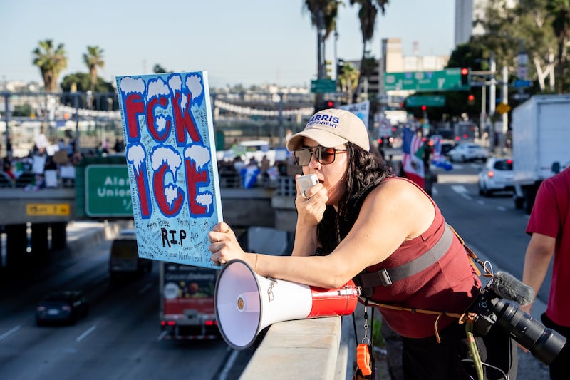 A woman in sunglasses and a beige baseball hat printed "Harris President" holds a canvas reading "Fck ICE" and "RIP" surrounded by small print of names of people who face injustice from immigration law enforcement.