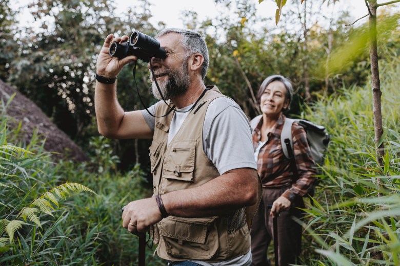 A man and woman standing amid brush, the man is looking through binoculars