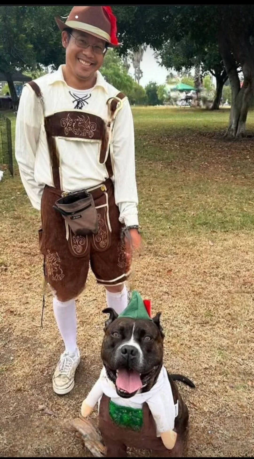 A man and a dog both wear lederhosen.