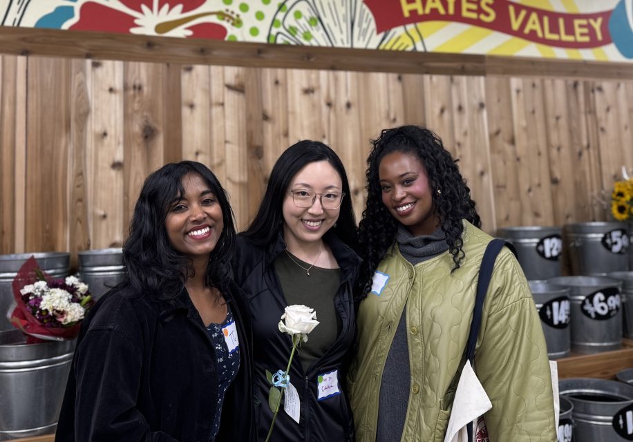Three women stand together and smile in front of a wooden wall with metal flower buckets in a store labeled "Hayes Valley," celebrating Valentine's Day. One woman holds a white rose.